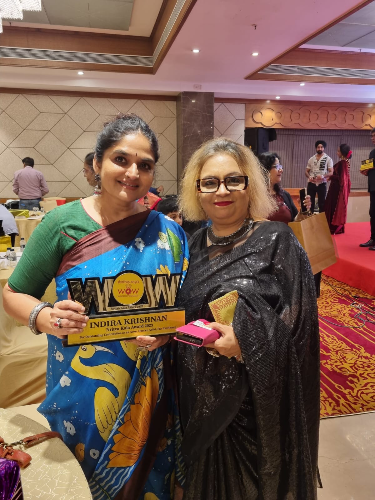 Two women wearing Kalamkari sarees including a blue and yellow batik saree by Kawaii at an indoor event