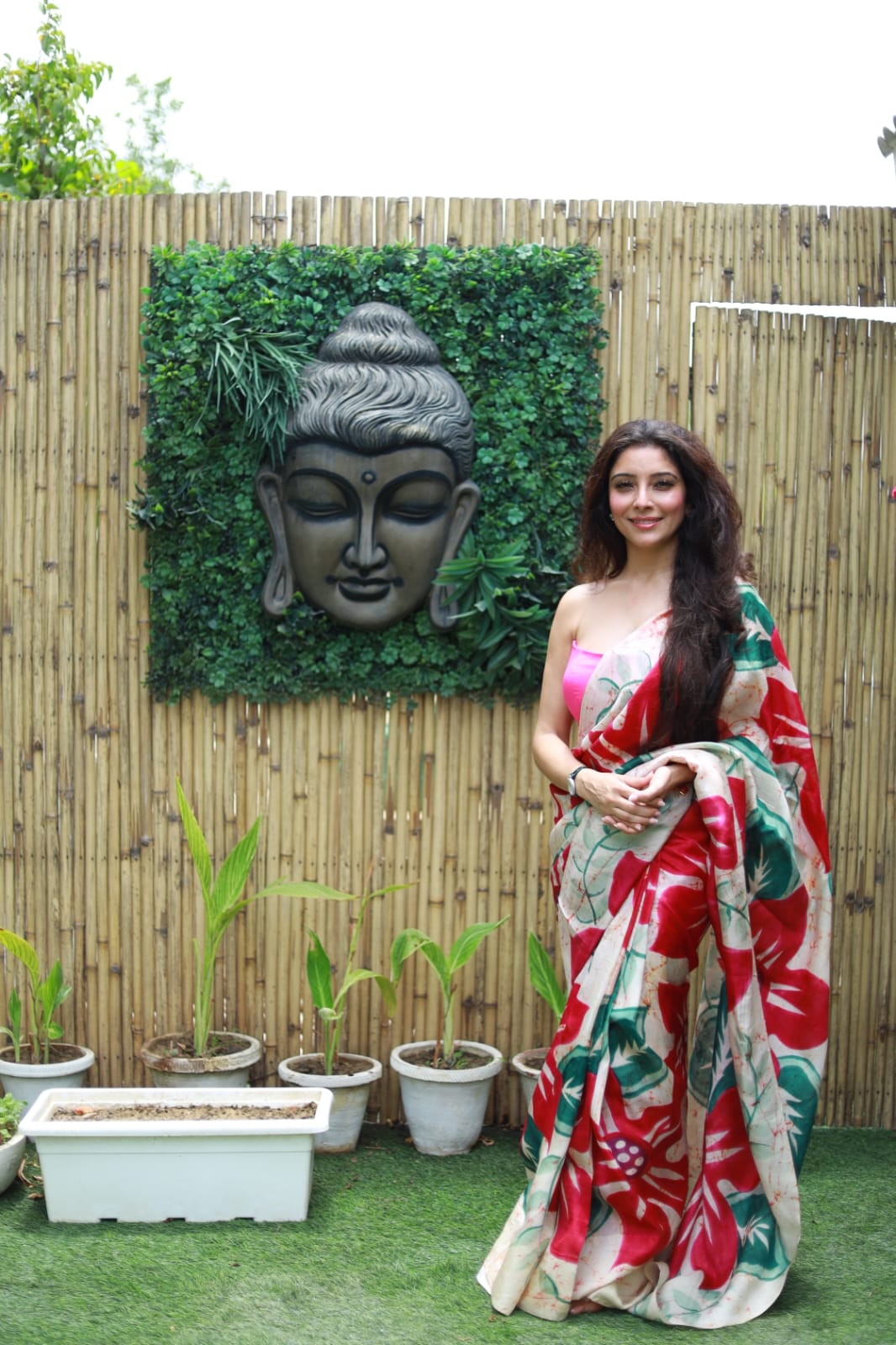 Woman wearing Kawaii batik saree with red and green floral patterns standing outdoors near a decorative Buddha wall art