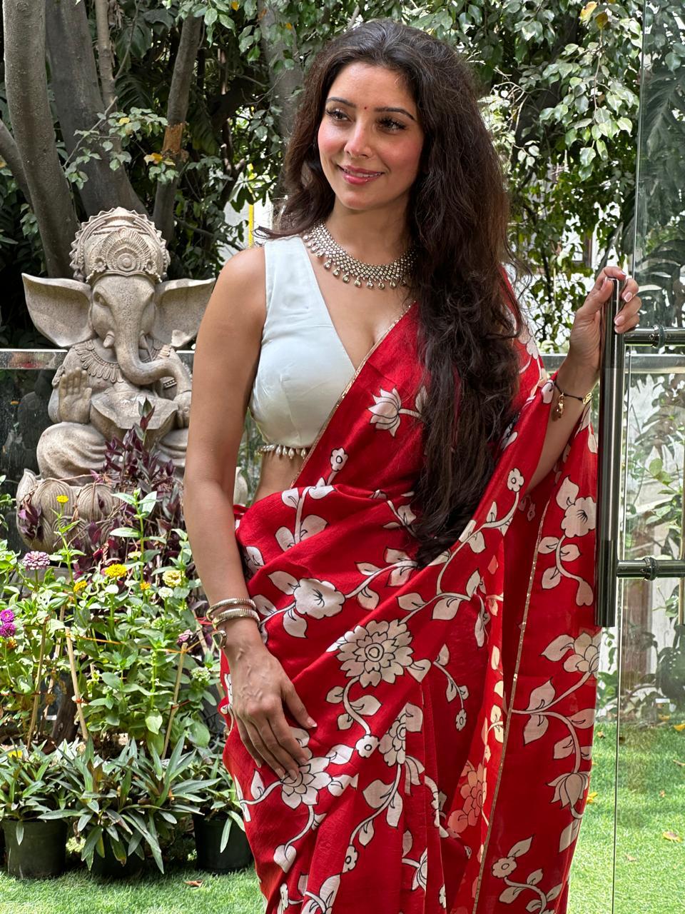 Woman wearing a red kalamkari saree with white floral patterns standing outdoors near plants and a statue