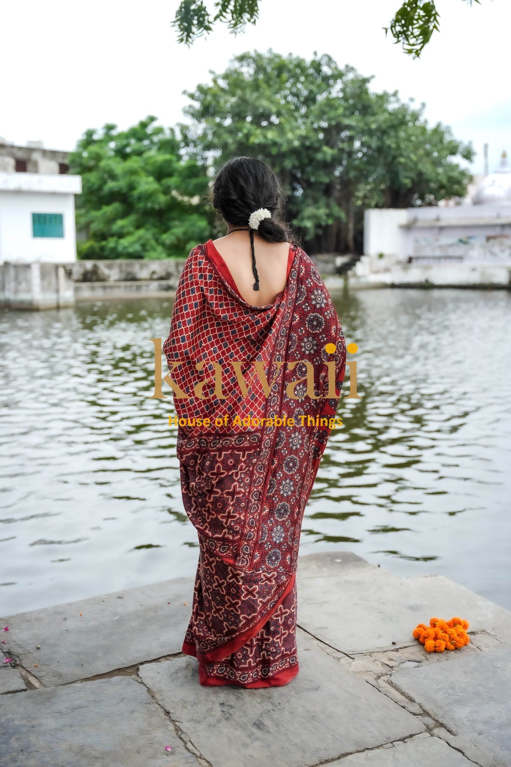 Woman wearing red patterned ajrakh saree standing by a riverside with greenery and buildings in the background