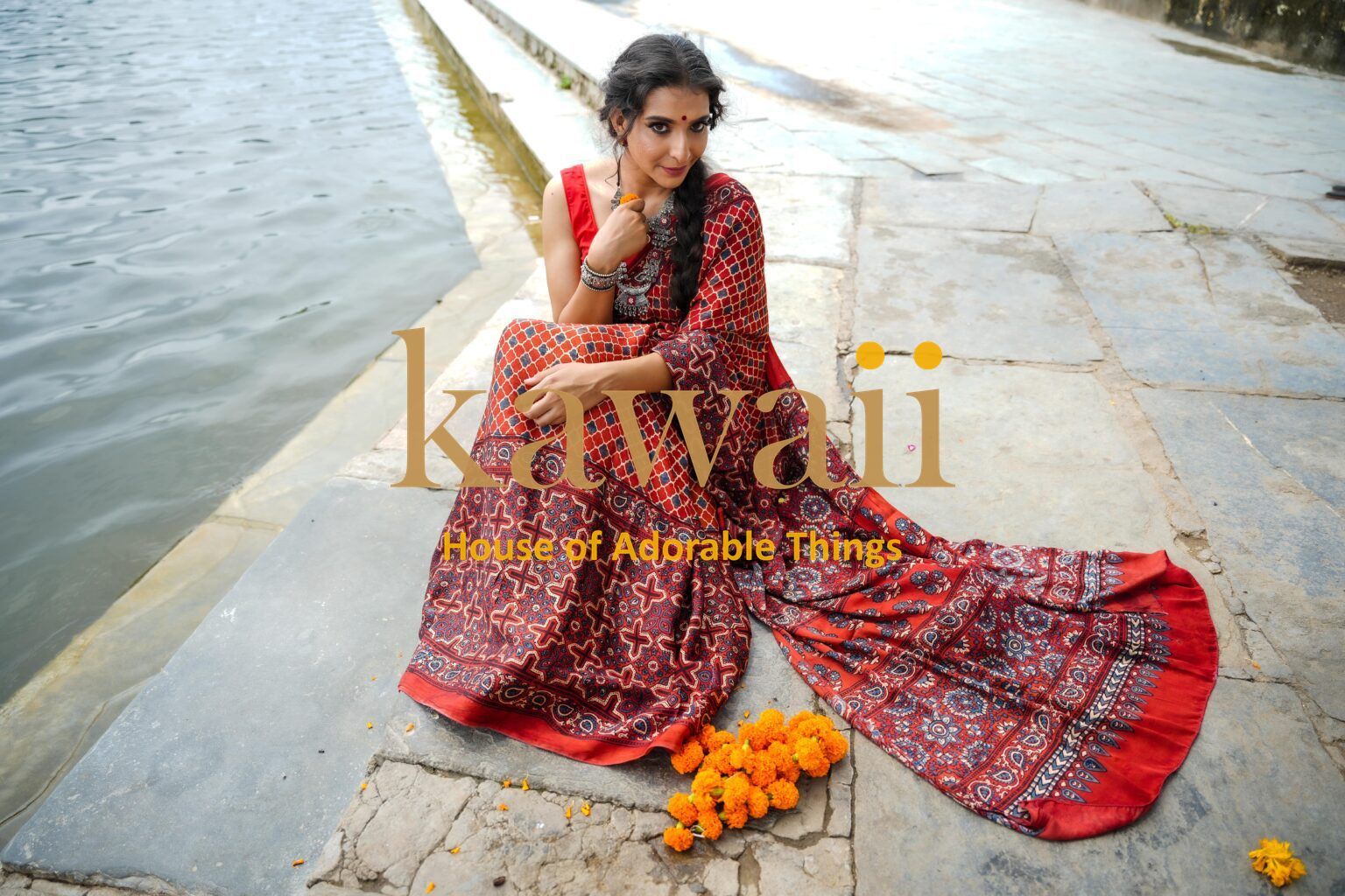 Woman wearing red patterned ajrakh saree sitting by water with orange flowers on stone steps