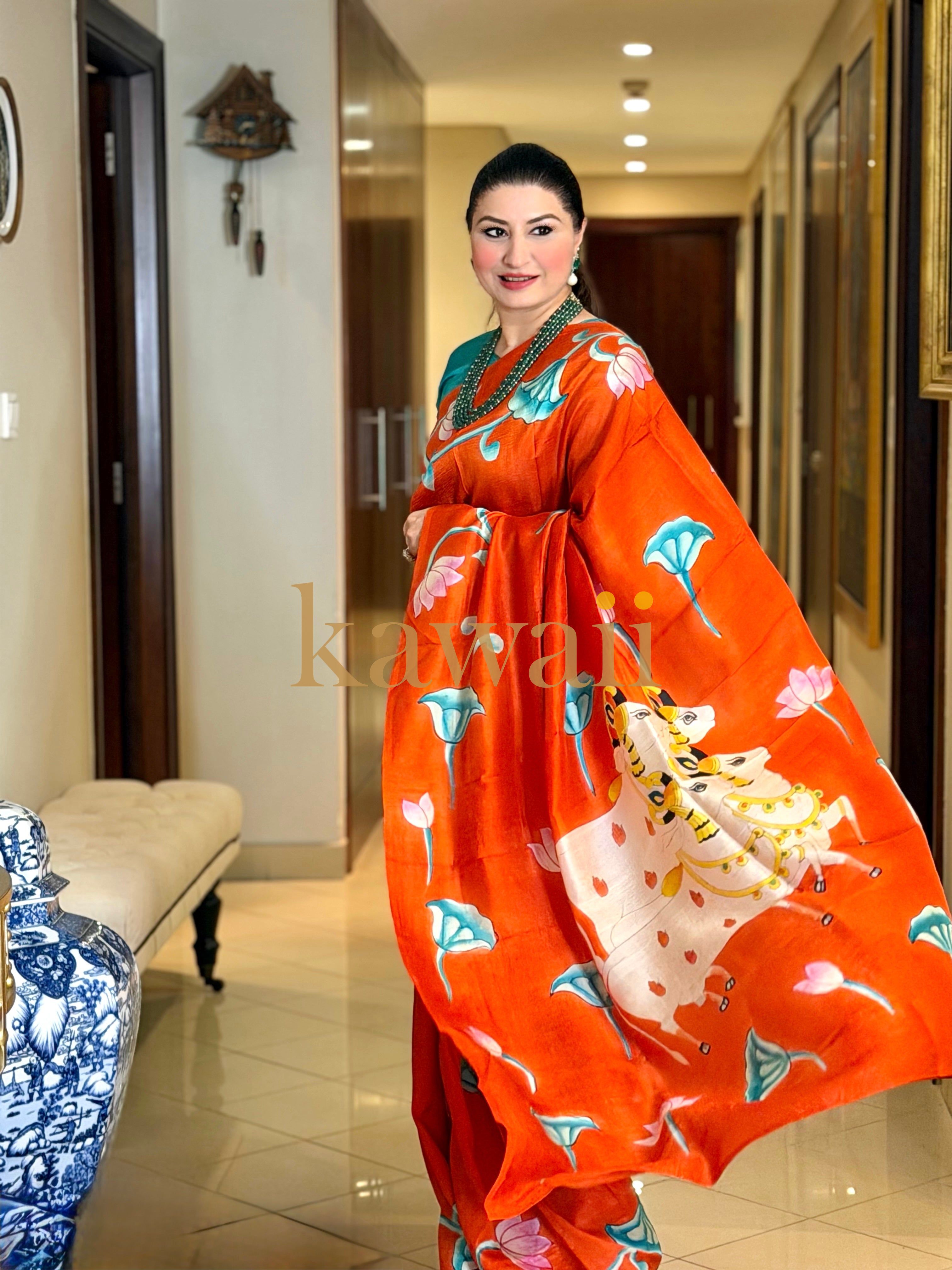 Stunning pichwai saree in vibrant orange with intricate floral designs, worn elegantly in a stylish home setting