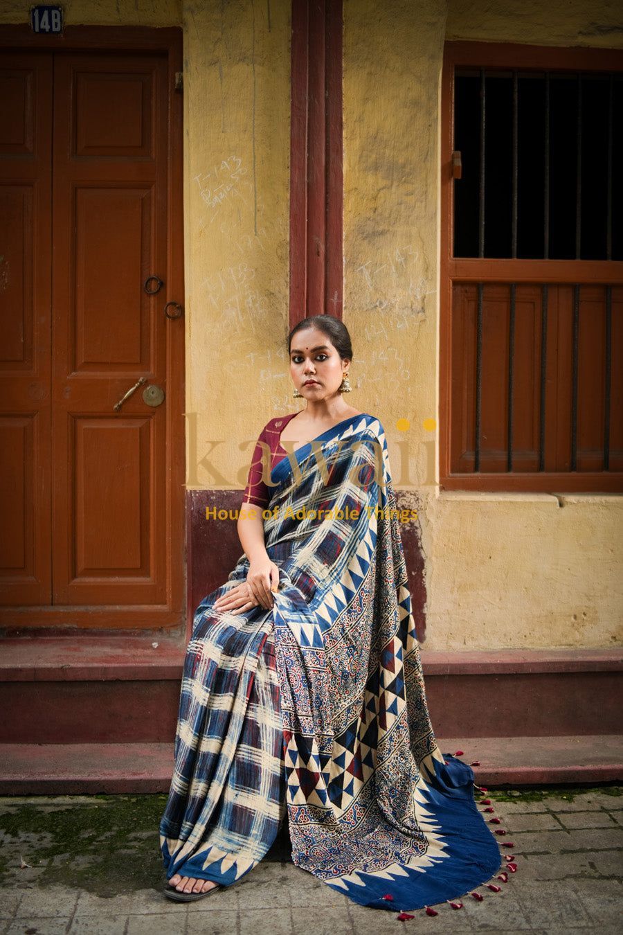 Woman wearing a traditional blue and beige ajrakh saree sitting outdoors by a rustic door