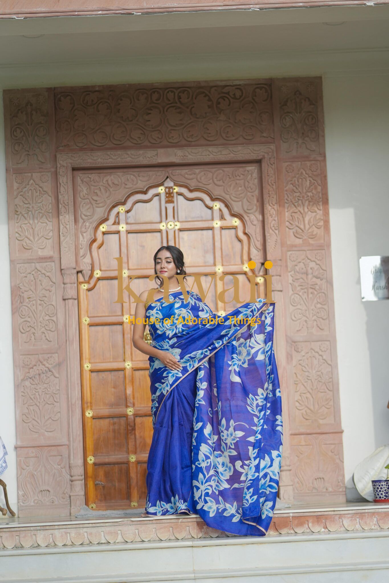 Woman wearing a blue batik saree with white floral patterns standing in front of a carved wooden door