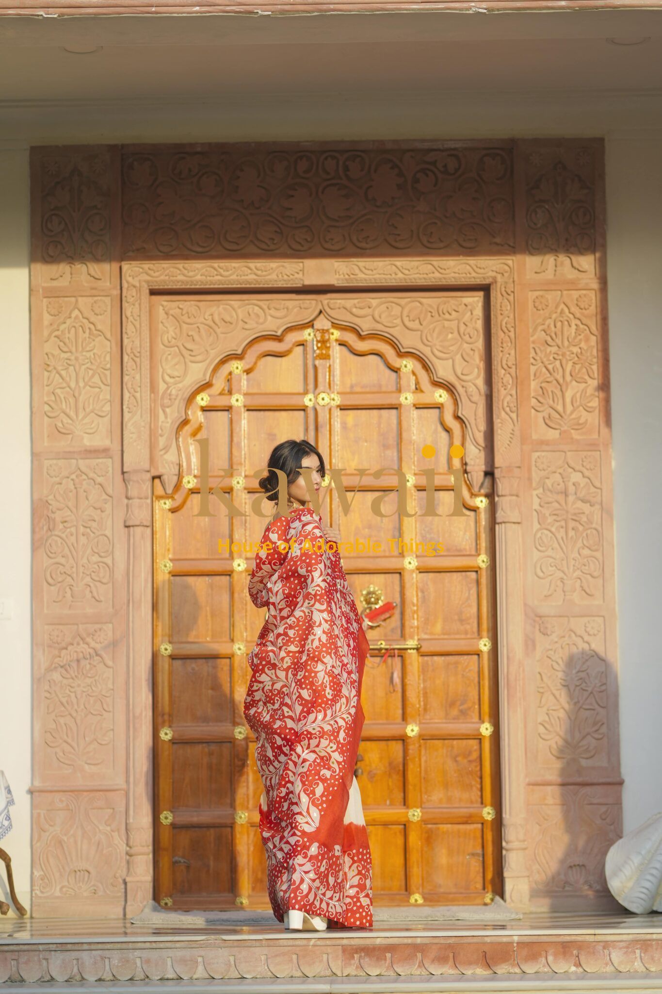 Woman wearing a red and white batik saree standing in front of a traditional carved wooden door