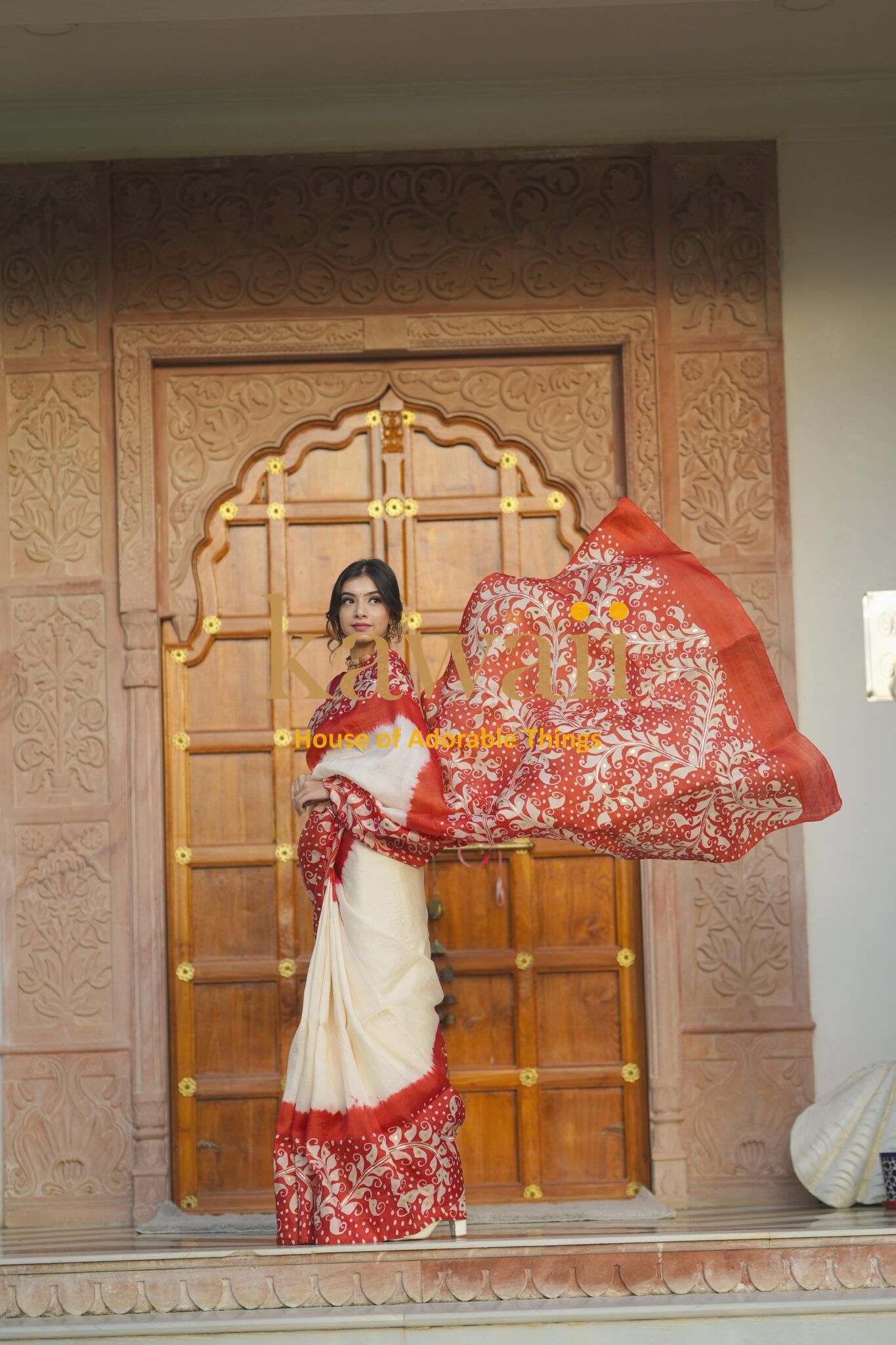 Kawaii batik saree in white and red with intricate floral patterns worn by a woman in front of a wooden carved door