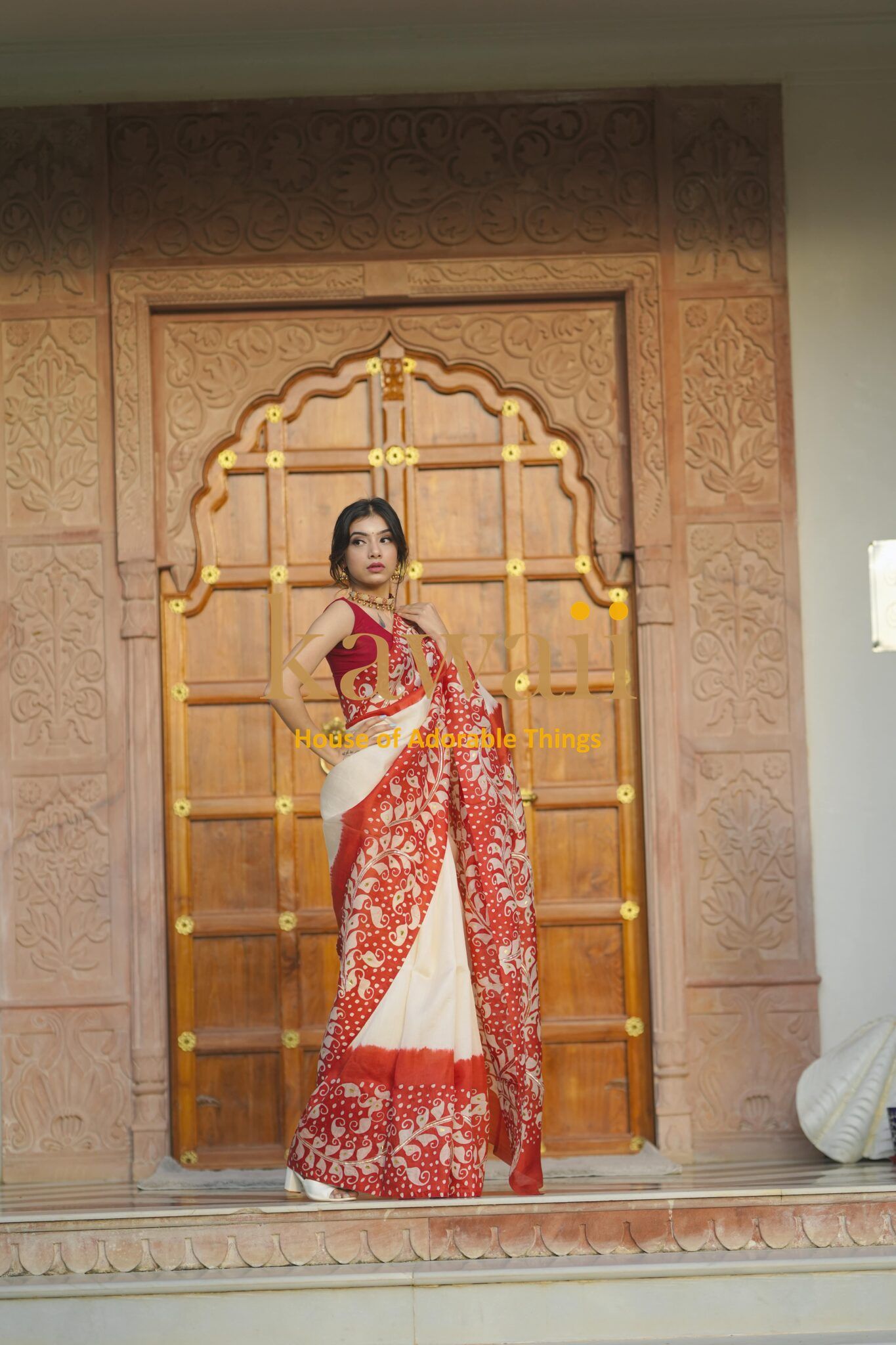 Kawaii batik saree in red and white worn by model standing in front of ornate wooden door