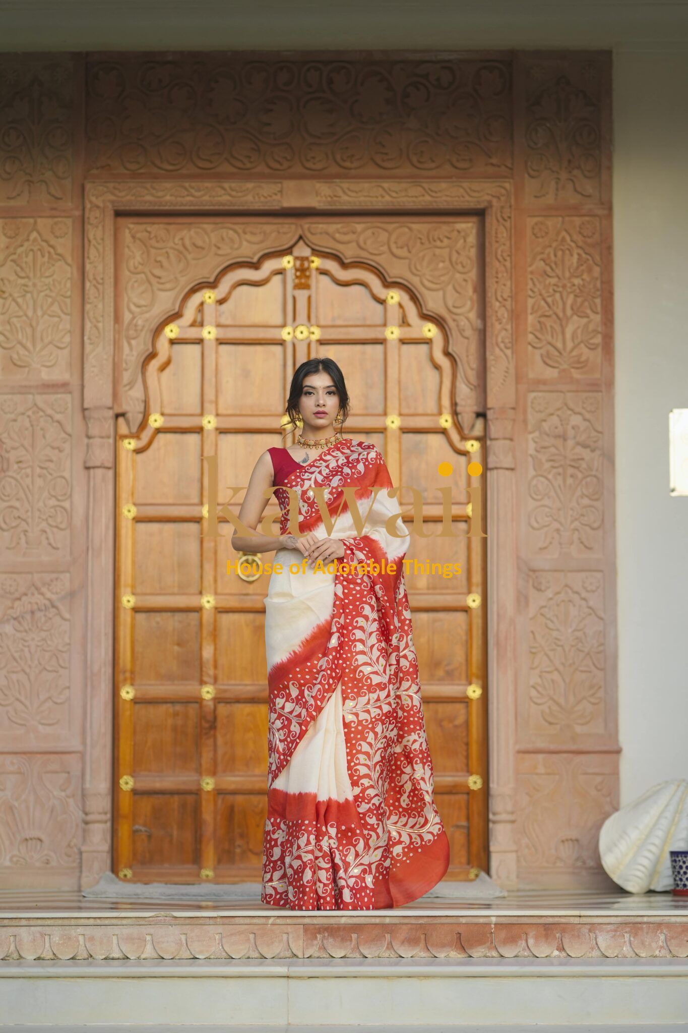 Kawaii batik saree in red and white with intricate floral patterns worn by a woman standing in front of a carved wooden door