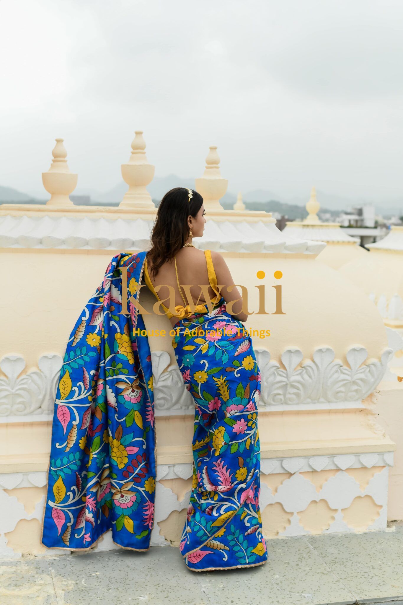 Woman wearing vibrant blue kalamkari saree with floral and bird motifs by Kawaii standing near ornate cream wall