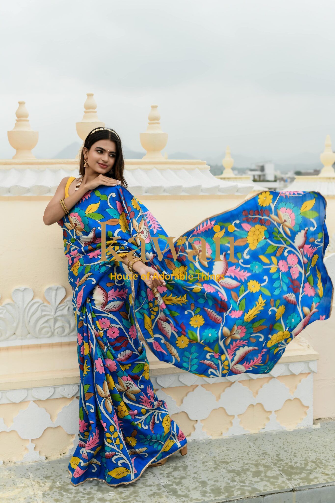 Woman wearing vibrant blue kalamkari saree with floral patterns standing outdoors by decorative wall