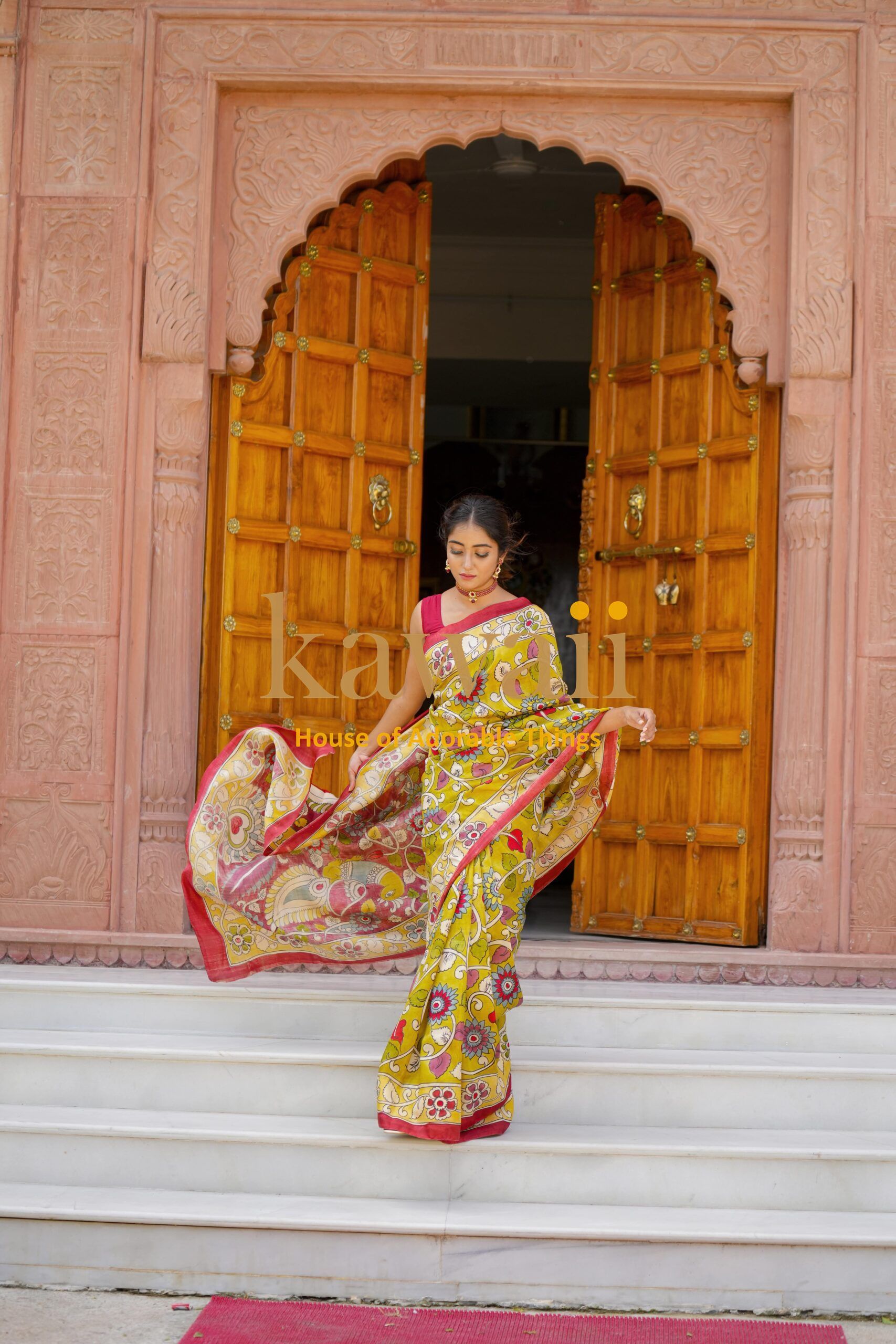 Woman wearing a yellow and red floral kalamkari saree standing in front of ornate wooden doors