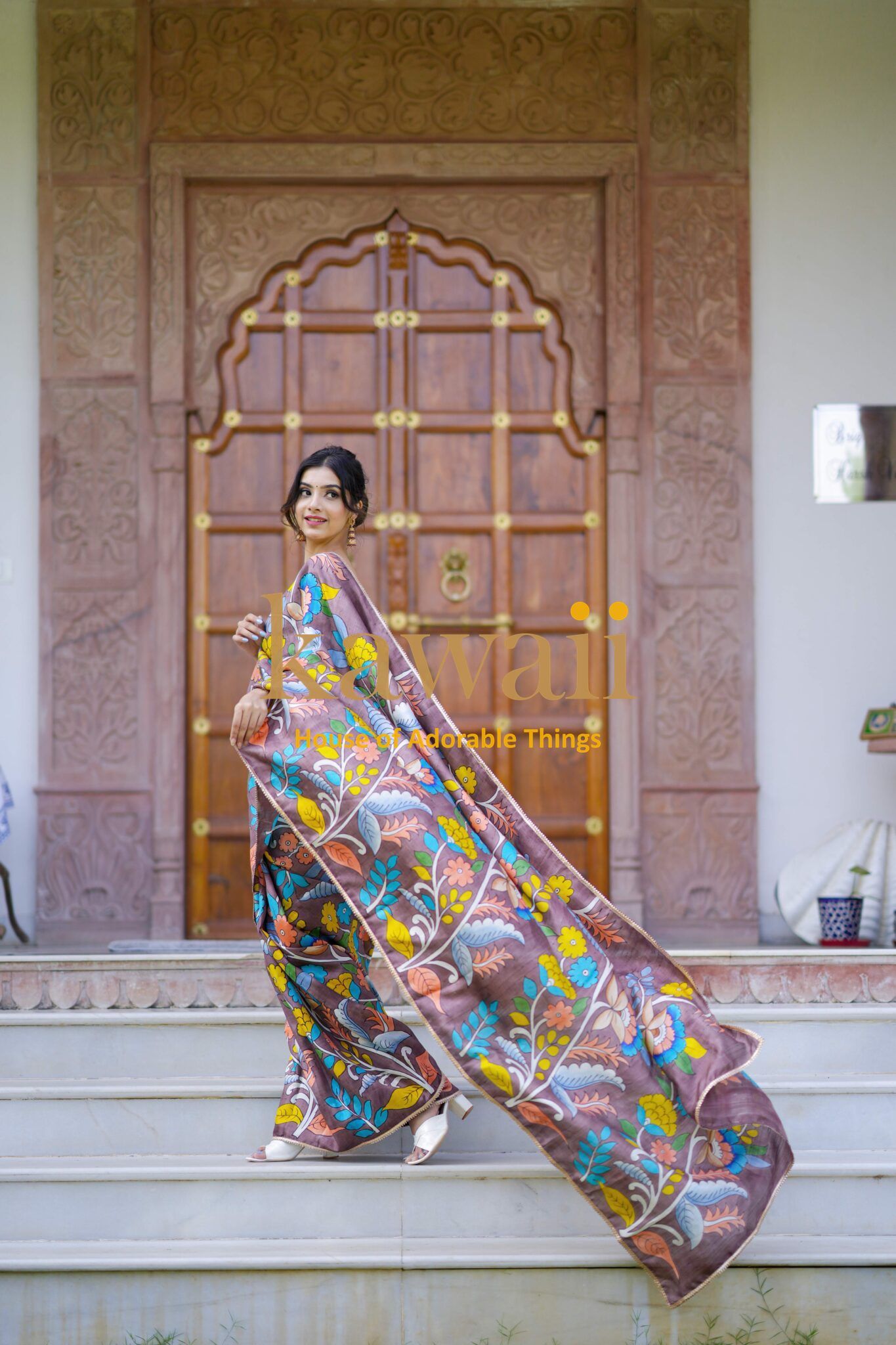 Woman wearing colorful Kawaii kalamkari saree with floral patterns standing on steps in front of ornate wooden door