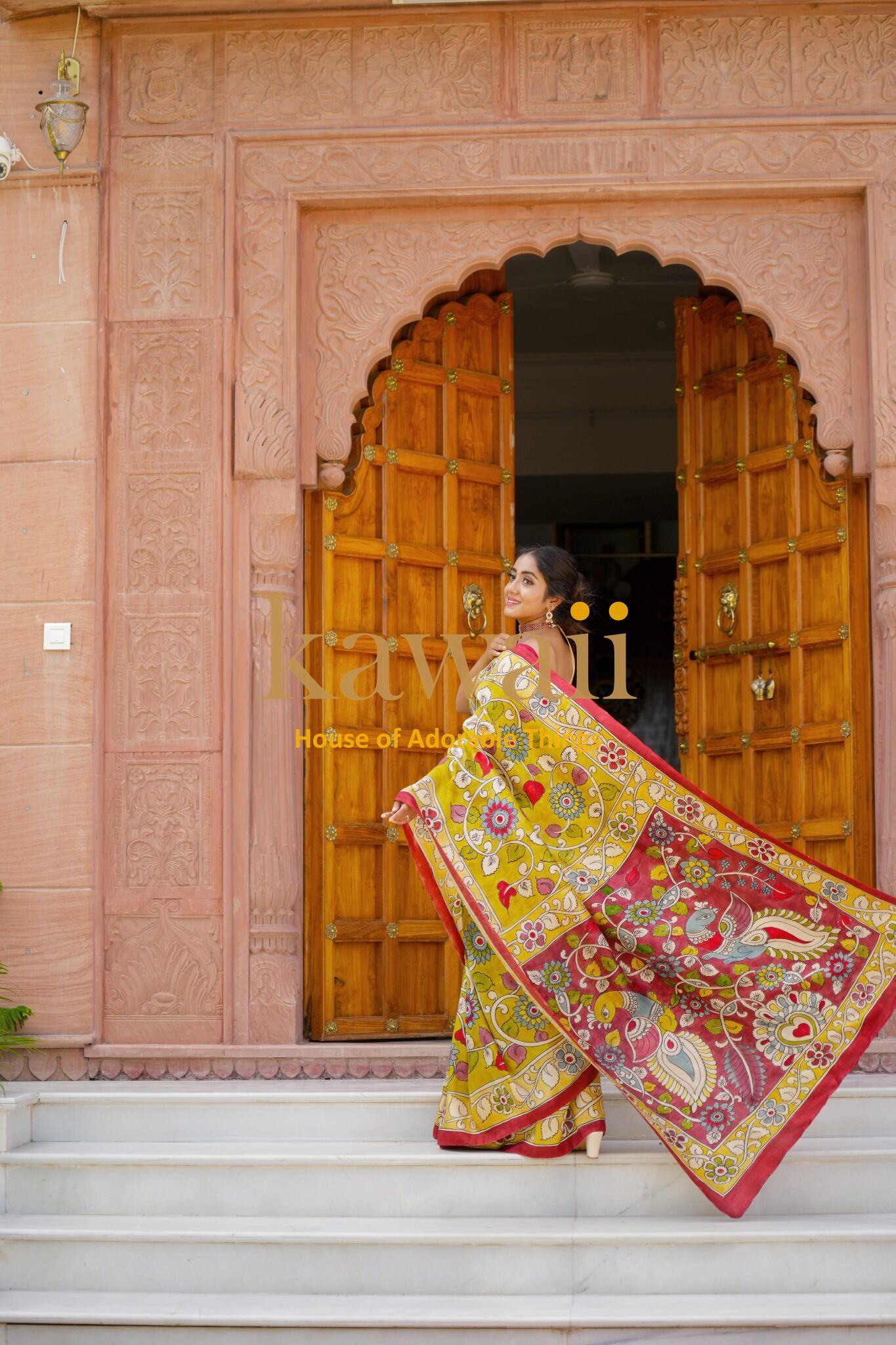 Woman wearing vibrant kalamkari saree with intricate floral and peacock designs standing by ornate wooden door