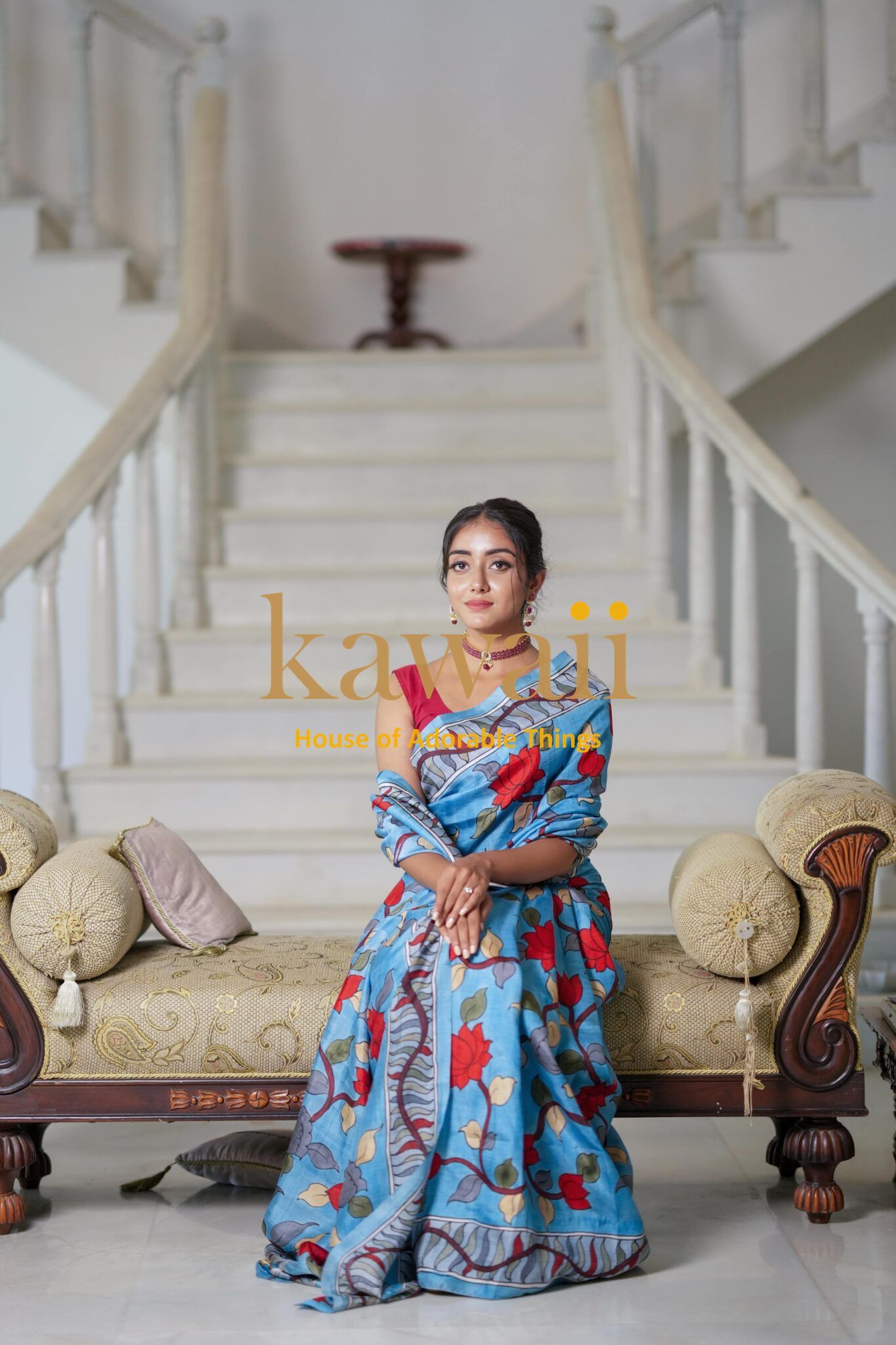 Woman wearing a blue floral kalamkari saree sitting on a beige cushioned sofa with staircase background