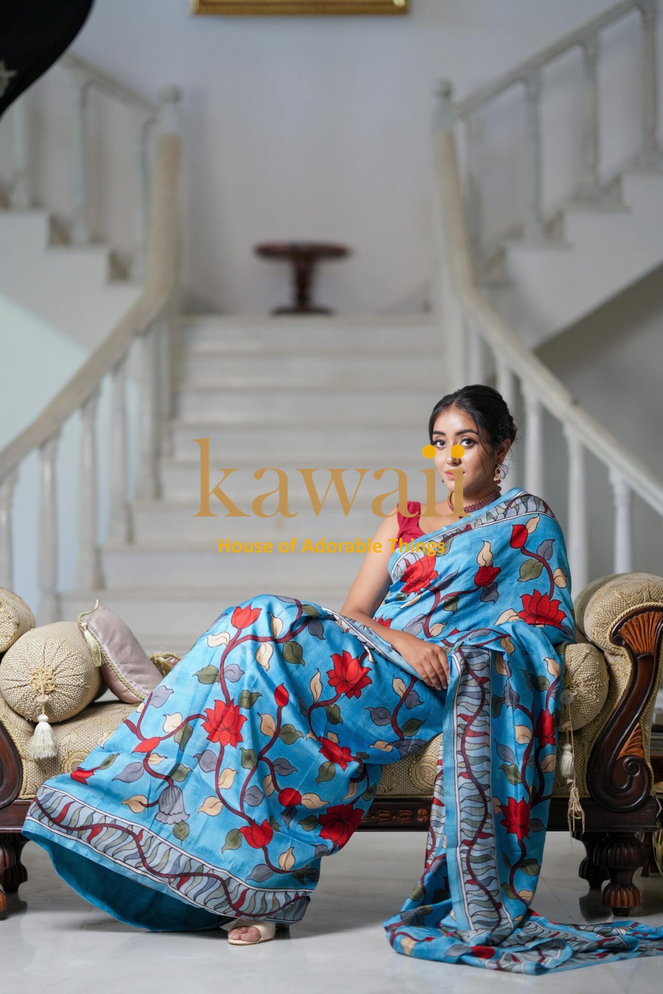 Woman wearing a blue and red floral design kalamkari saree sitting on an antique sofa indoors