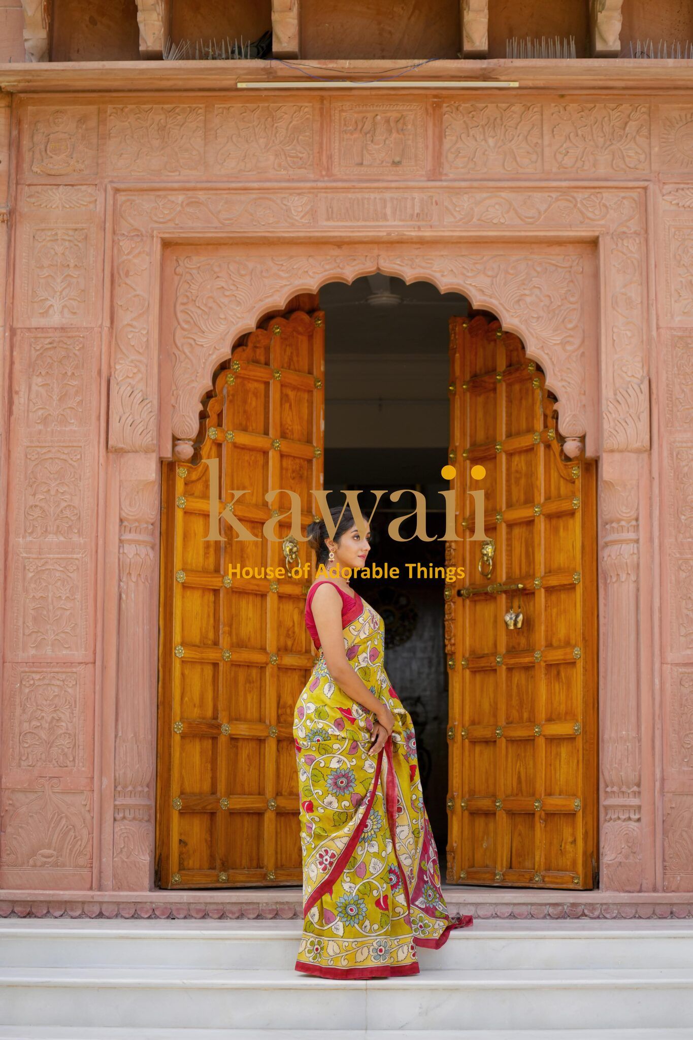 Woman wearing a yellow and pink kalamkari saree standing in front of wooden doors with intricate architecture