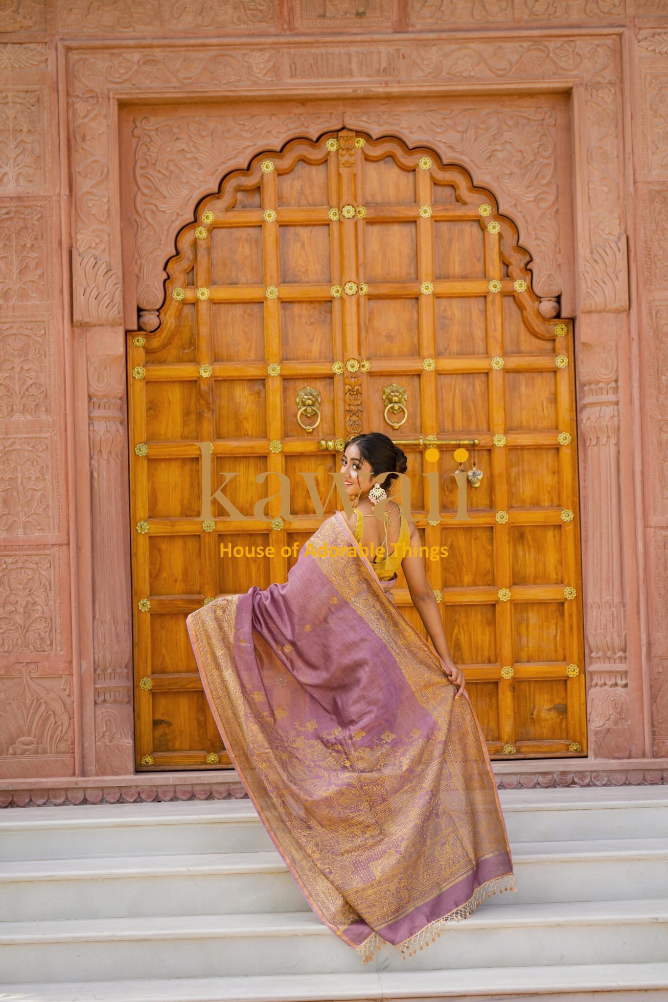 Woman wearing a purple and gold madhubani saree standing in front of an ornate wooden door