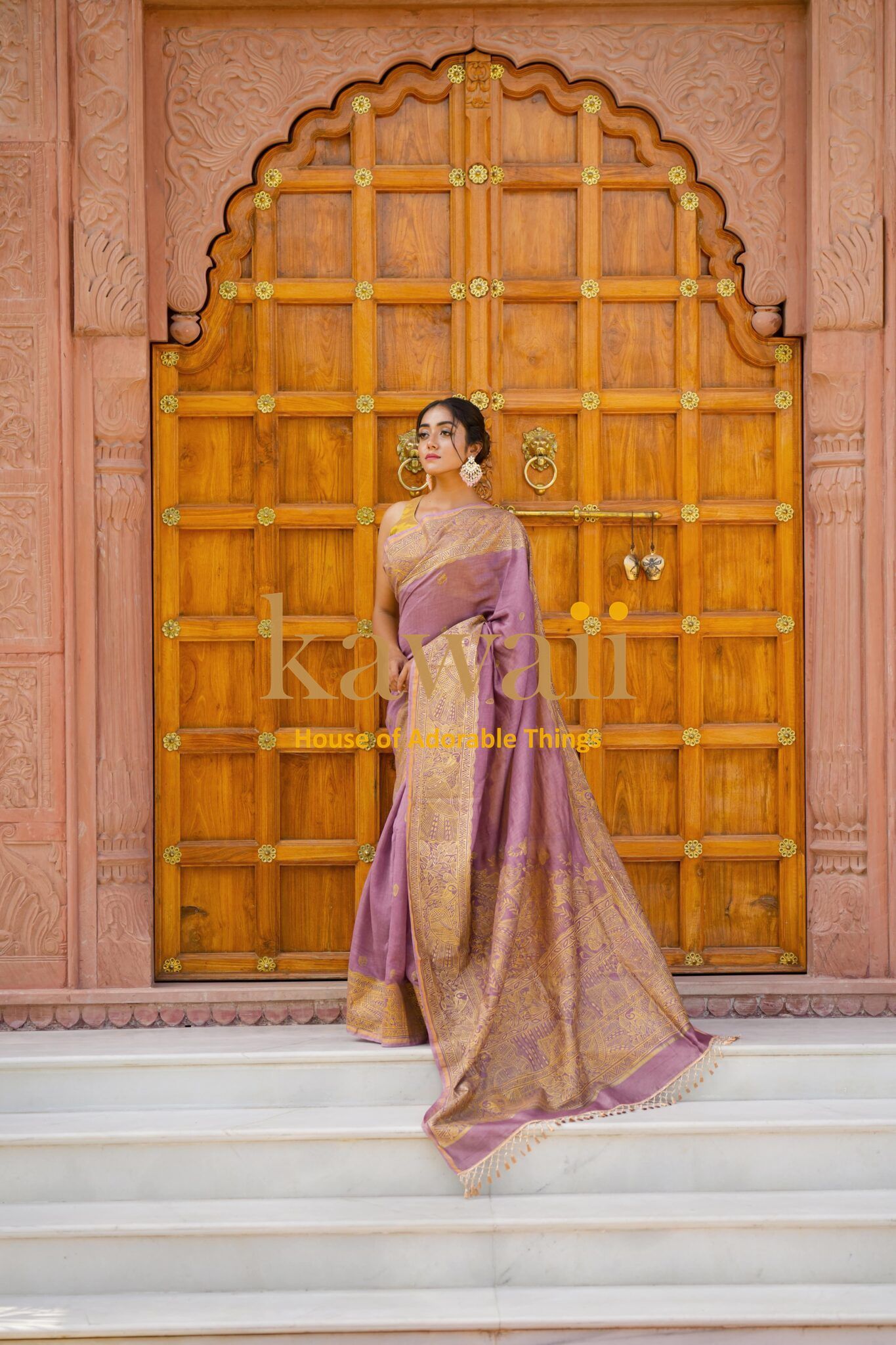 Elegant woman wearing a purple and gold madhubani saree standing in front of a carved wooden door