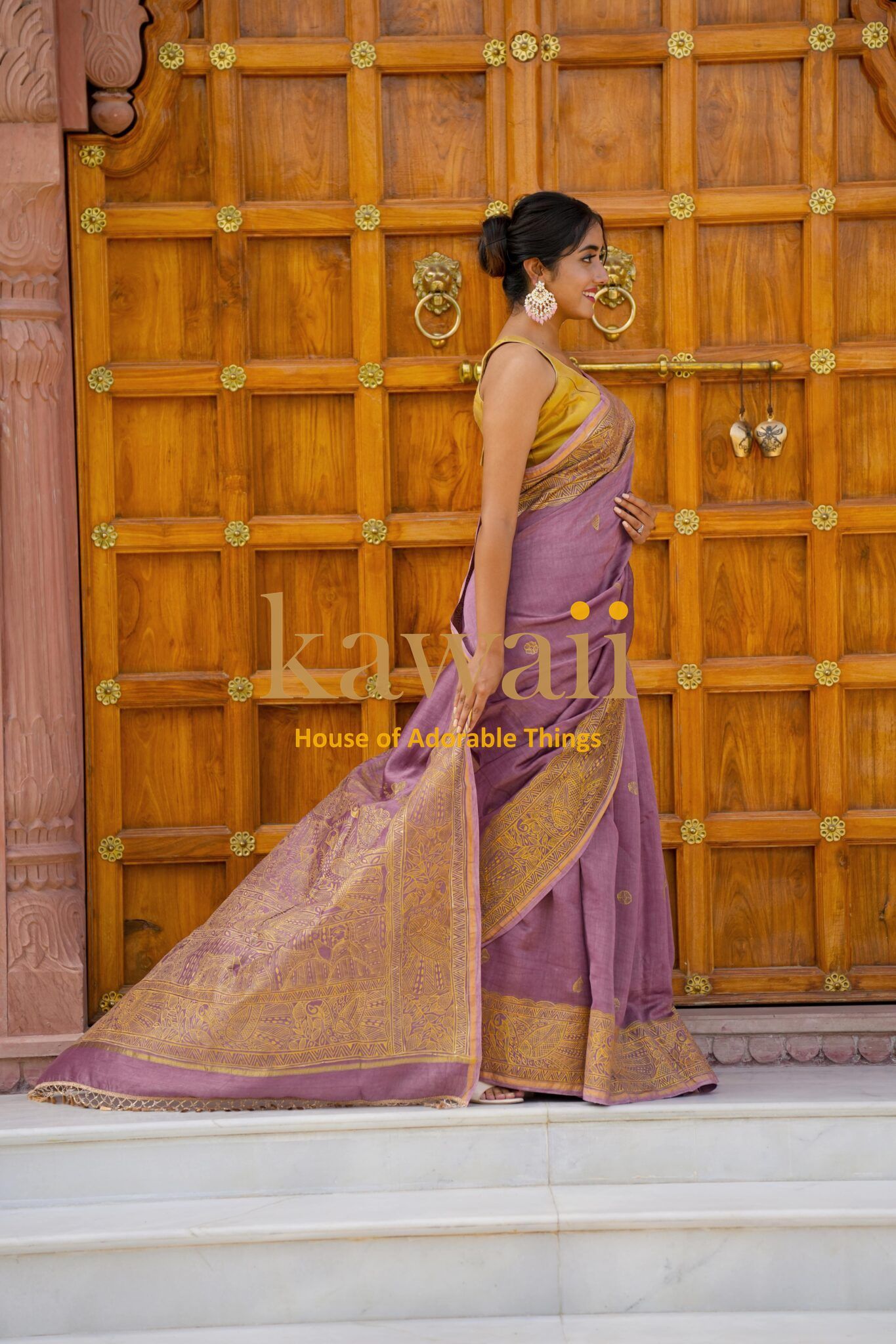Kawaii Madhubani saree in purple with gold intricate patterns draped on woman in front of wooden door