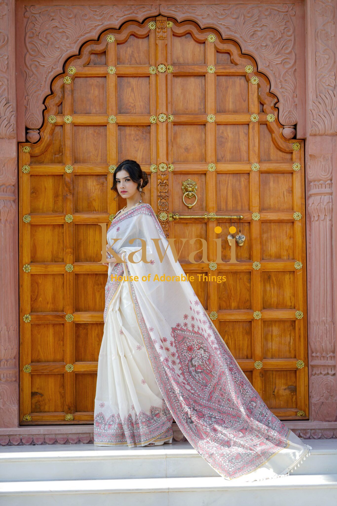 Woman wearing elegant white and pink Madhubani saree standing in front of a wooden carved door