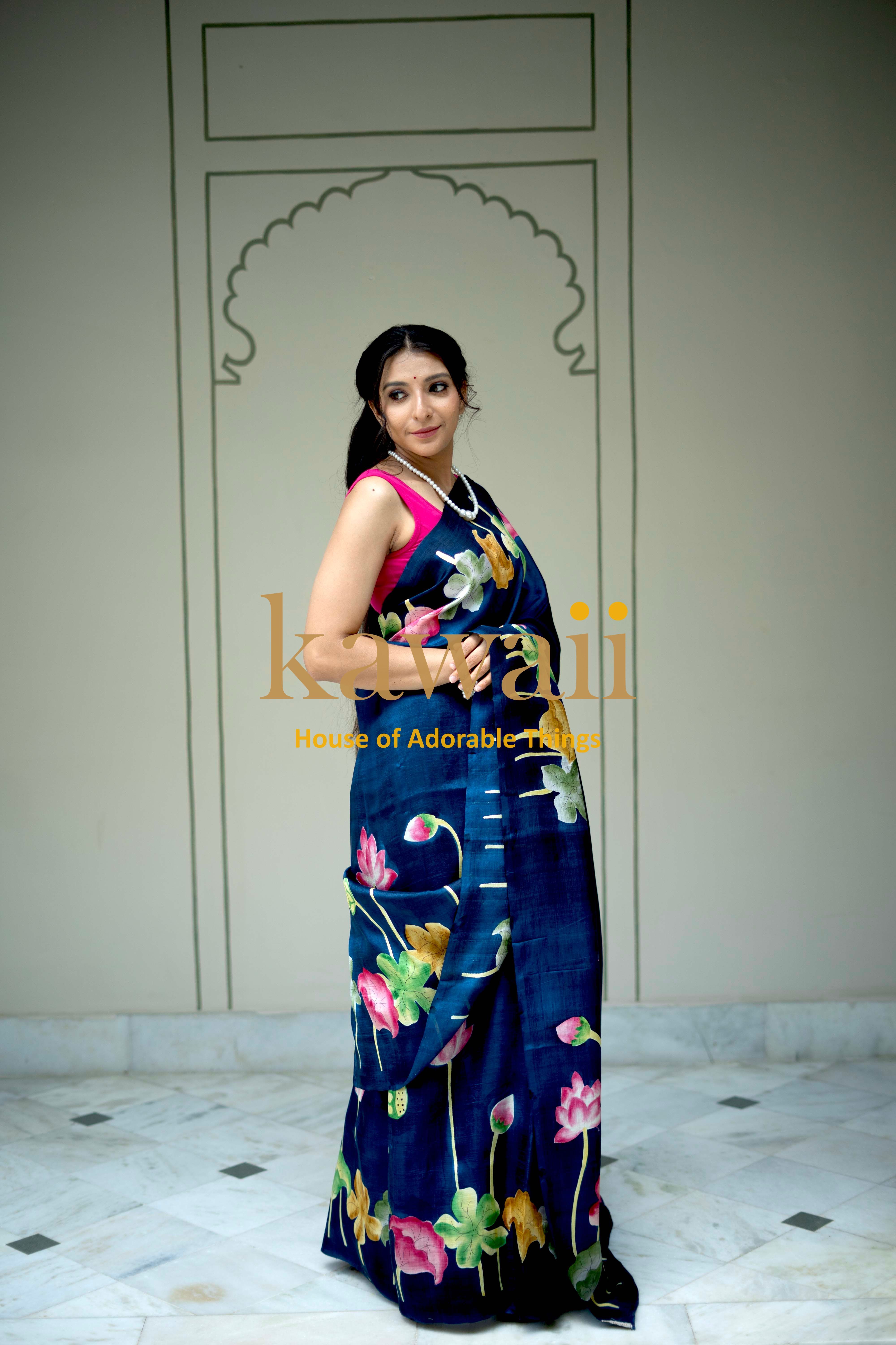 Woman wearing a blue pichwai saree with floral patterns standing indoors with decorative wall