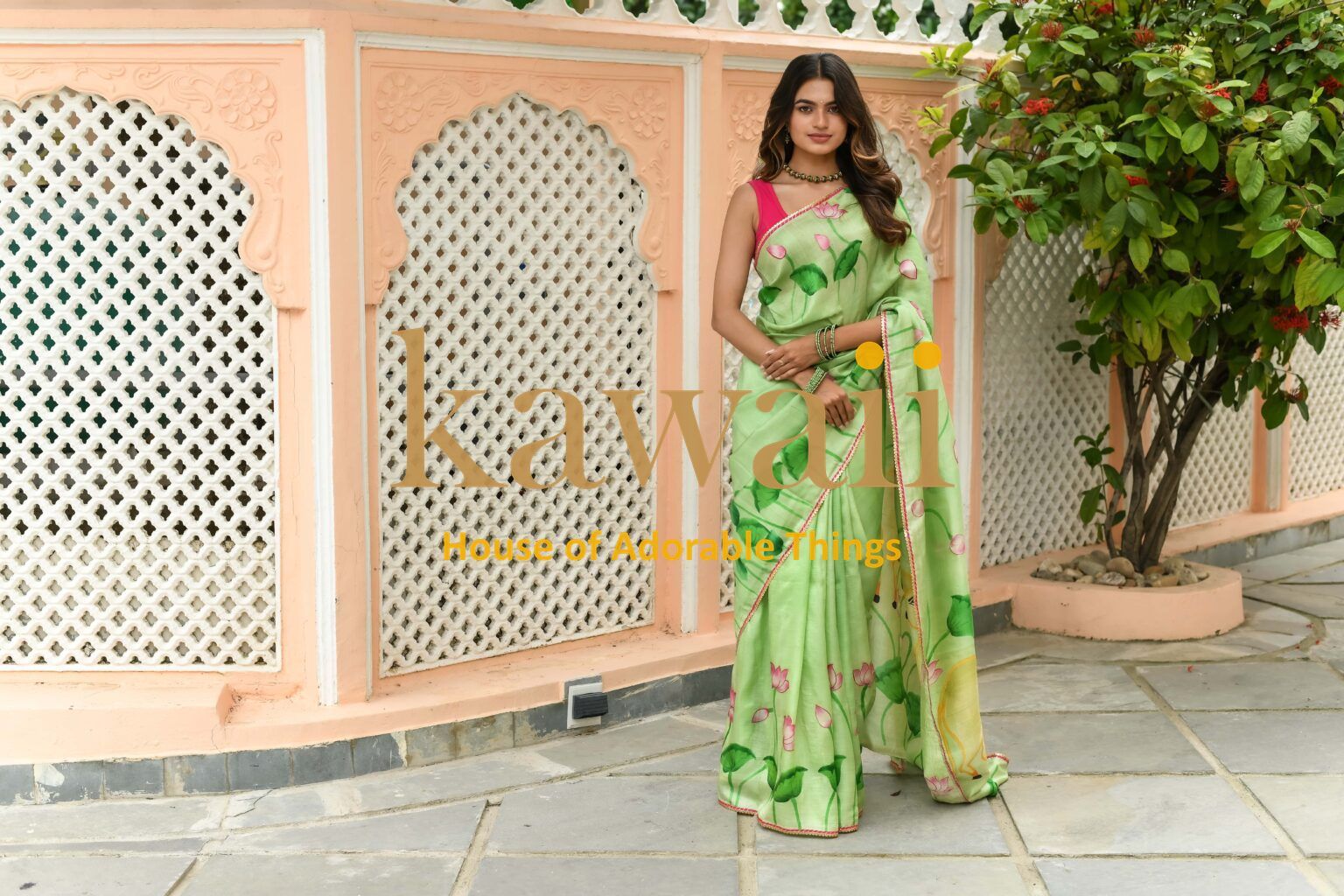 Model wearing a green floral pichwai saree by Kawaii standing outdoors near a peach-colored architectural wall