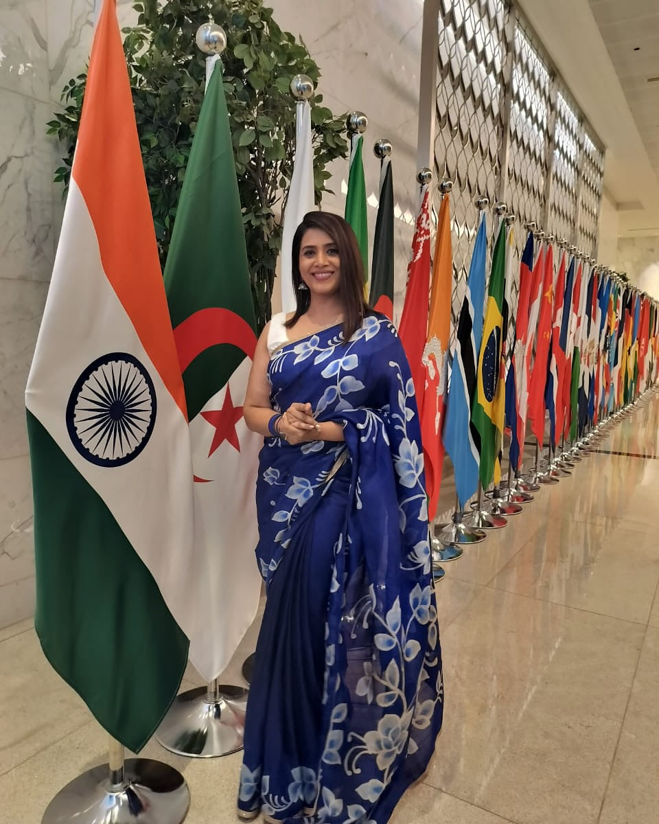 Woman wearing a blue floral batik saree standing beside international flags in a hallway