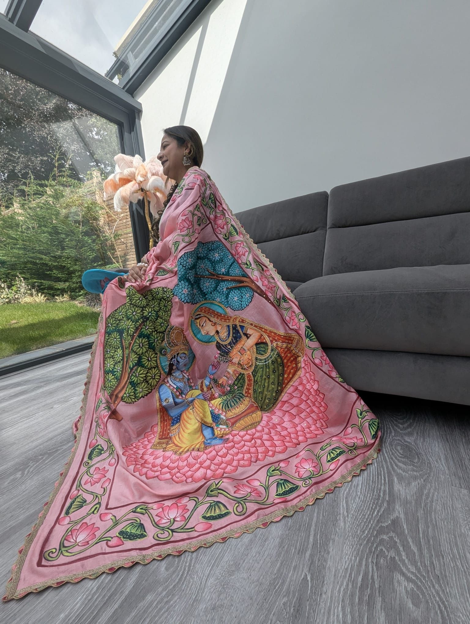 Woman wearing pink pichwai saree with detailed traditional Indian artwork sitting near a window