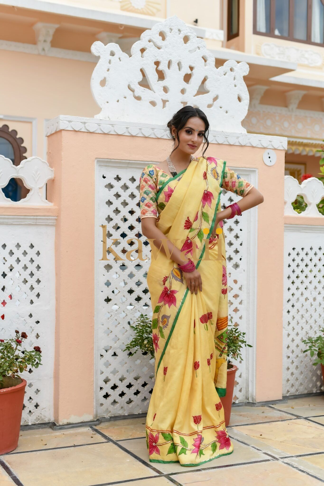 Woman wearing a yellow floral pichwai saree standing outdoors near decorative white lattice wall