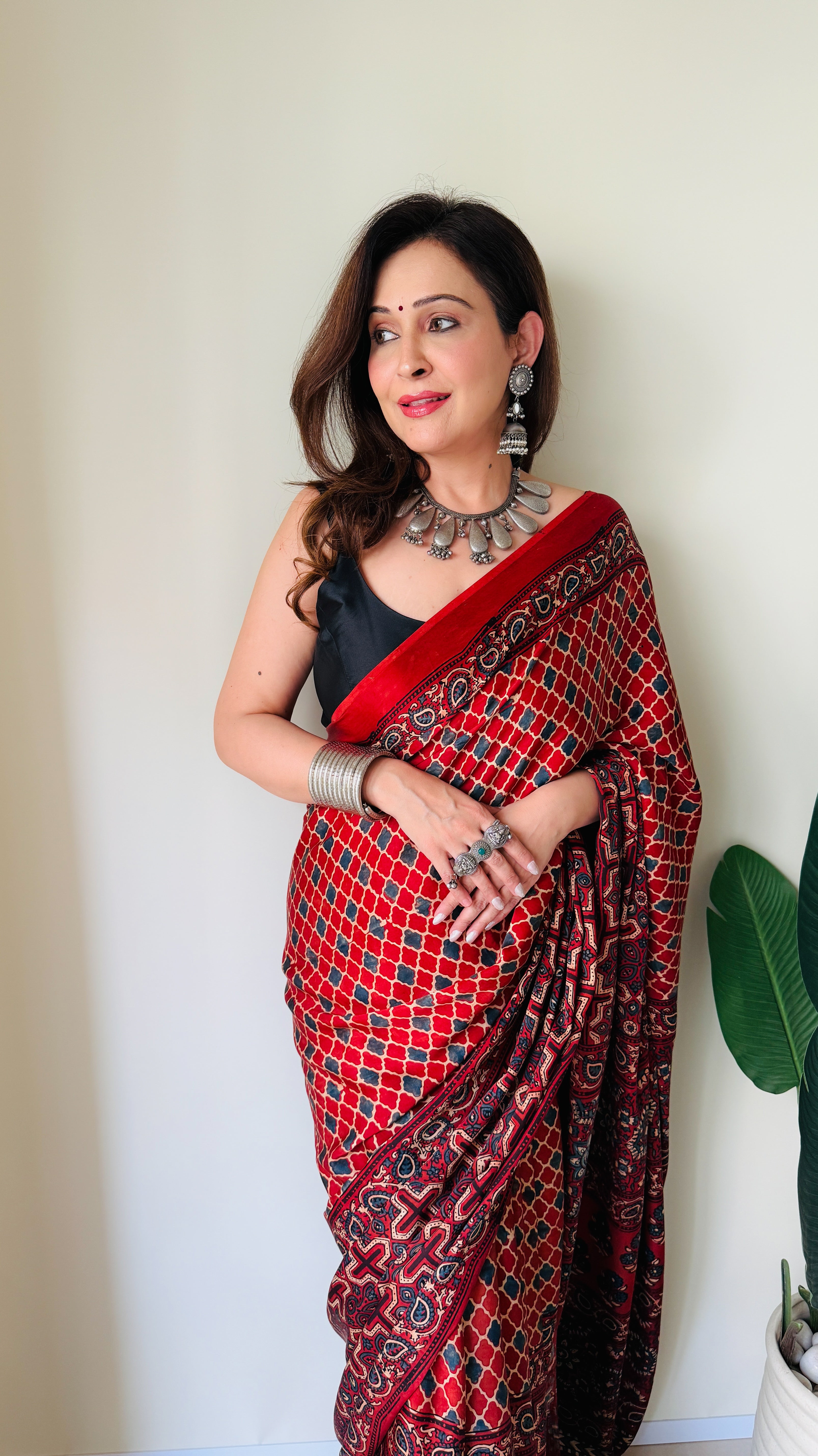 Woman wearing a red and black patterned ajrakh saree with silver jewelry standing against a white wall